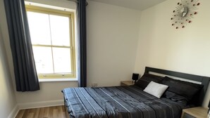 Quarto com cama, mesas de cabeceira, janela com cortinas, relógio de parede e luminária no teto.