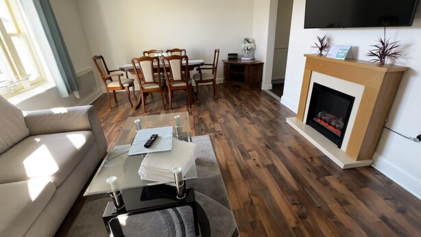 Uma sala de estar com sofá, mesa de centro de vidro com controle remoto, lareira e um espaço de refeições com cadeiras e mesa.