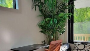 Quarto com cadeira de madeira, escrivaninha, uma planta grande em vaso, cama com colcha branca e cinza, e janela com vista para vegetação.