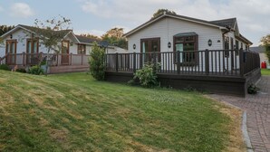 A row of white mobile homes with wooden decks and railings, set on a grassy slope with a clear sky above. - 27 Brocklehurst (Oakham)