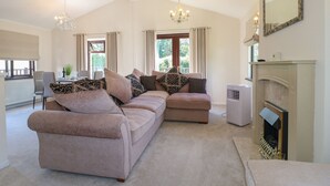 A spacious living room with a beige sofa set, a fireplace, and a chandelier. - 27 Brocklehurst (Oakham)