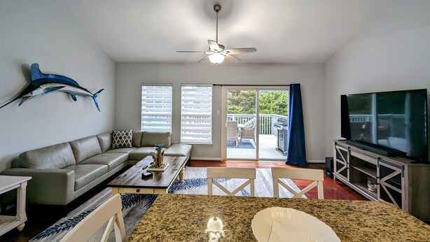 A living room with a sofa, coffee table, and a television. There is a ceiling fan, a large fish decoration on the wall, and a sliding glass door leading to an outdoor area with a grill.