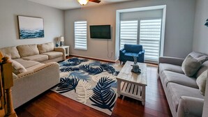 A living room with a ceiling fan, a large window with blinds, a television mounted on the wall, and a sofa.