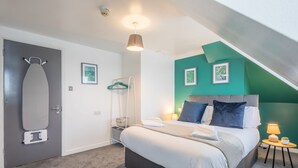 A bedroom with a bed, a hanging laundry rack, a door with an ironing board, and a small table with a lamp.
