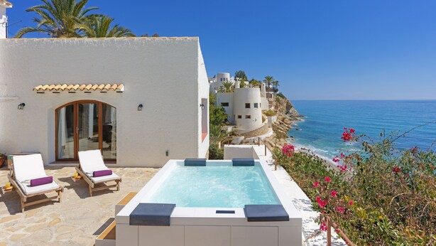A pool area with lounge chairs and a hot tub, overlooking a coastal landscape with buildings and palm trees.