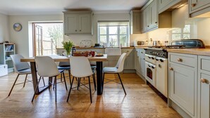 A modern kitchen with light-colored cabinets, a wooden countertop, and a copper pendant light. There is a dining table with chairs and a window providing natural light. - Walnut Cottage | Evenley Holiday Let | 2 Beds & Sleeps 4 (EVENLEY, NEAR BICESTER)
