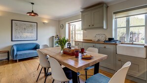 A cozy kitchen with a wooden table, chairs, and a blue sofa. There is a window with a view of the outside, and a map is hanging on the wall. - Walnut Cottage | Evenley Holiday Let | 2 Beds & Sleeps 4 (EVENLEY, NEAR BICESTER)