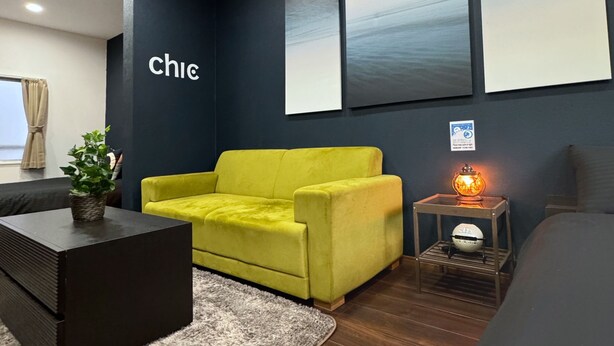 A modern living room with a yellow sofa, a black coffee table, a small side table with a lamp, and a bed in the background. - 【大阪No.1ロケーション】JR大阪駅、人気の梅田エリアから徒歩圏内。大阪観光地、USJ、難波、京都へ好アクセス！ (大阪市北区中津)