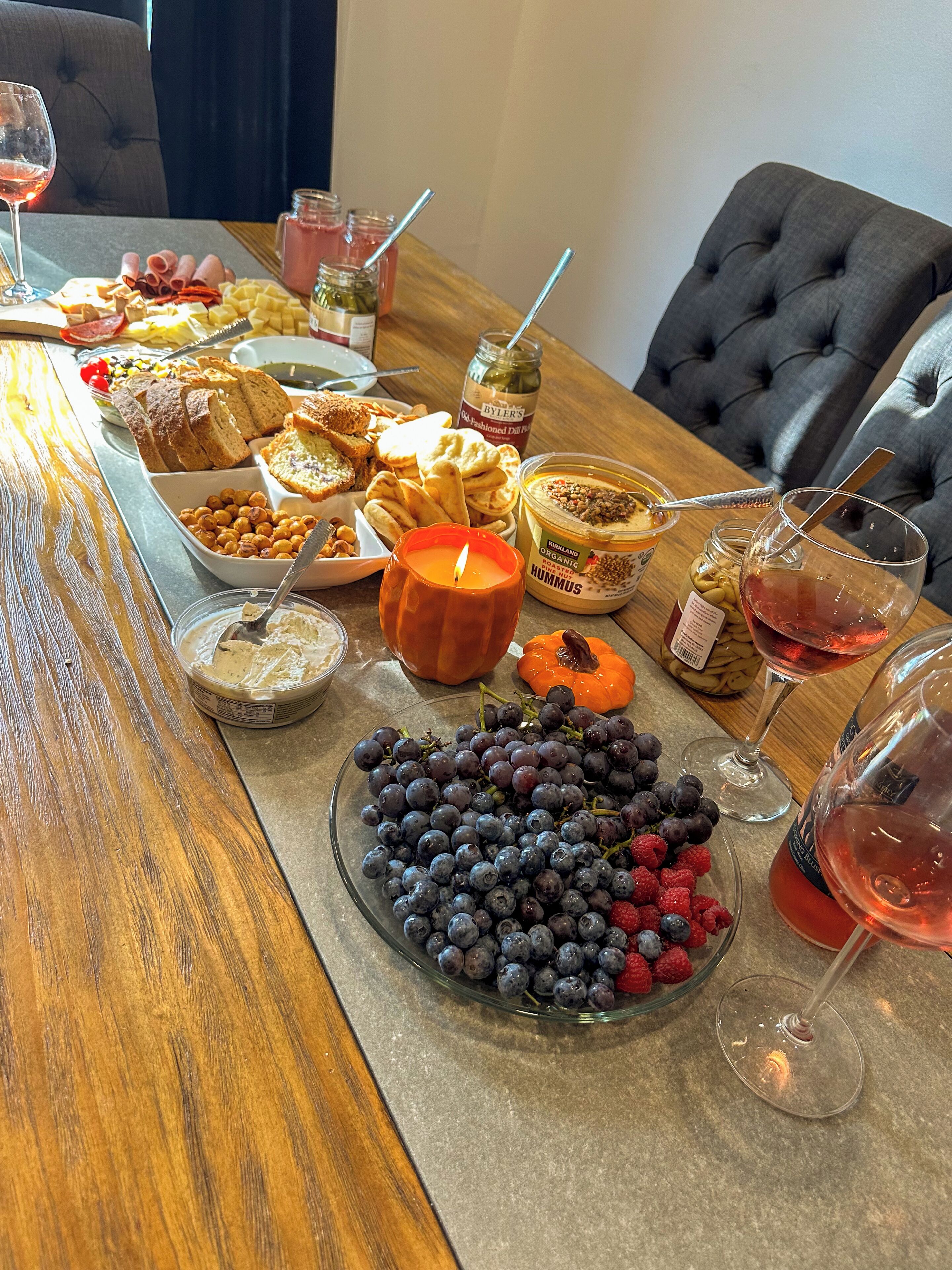 A dining table with various food items and drinks, including a bowl of grapes and raspberries, a candle, and jars of preserves.