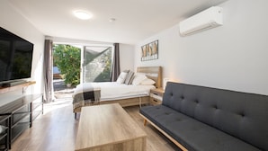 Una habitación moderna y luminosa con una cama, un sofá gris, una mesa de centro de madera y un televisor sobre un mueble.
