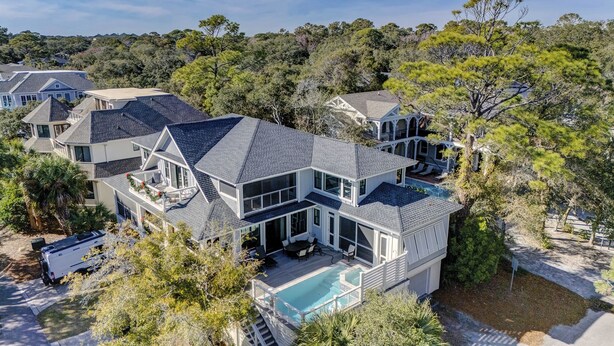 A large, multi-story house with a swimming pool, surrounded by dense trees and other houses.