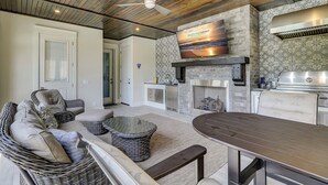 A spacious outdoor area with a stone fireplace, a flat-screen TV mounted above it, wicker furniture, and a ceiling fan.