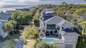 A modern two-story house with a large deck and a swimming pool, surrounded by lush greenery and a view of the beach.