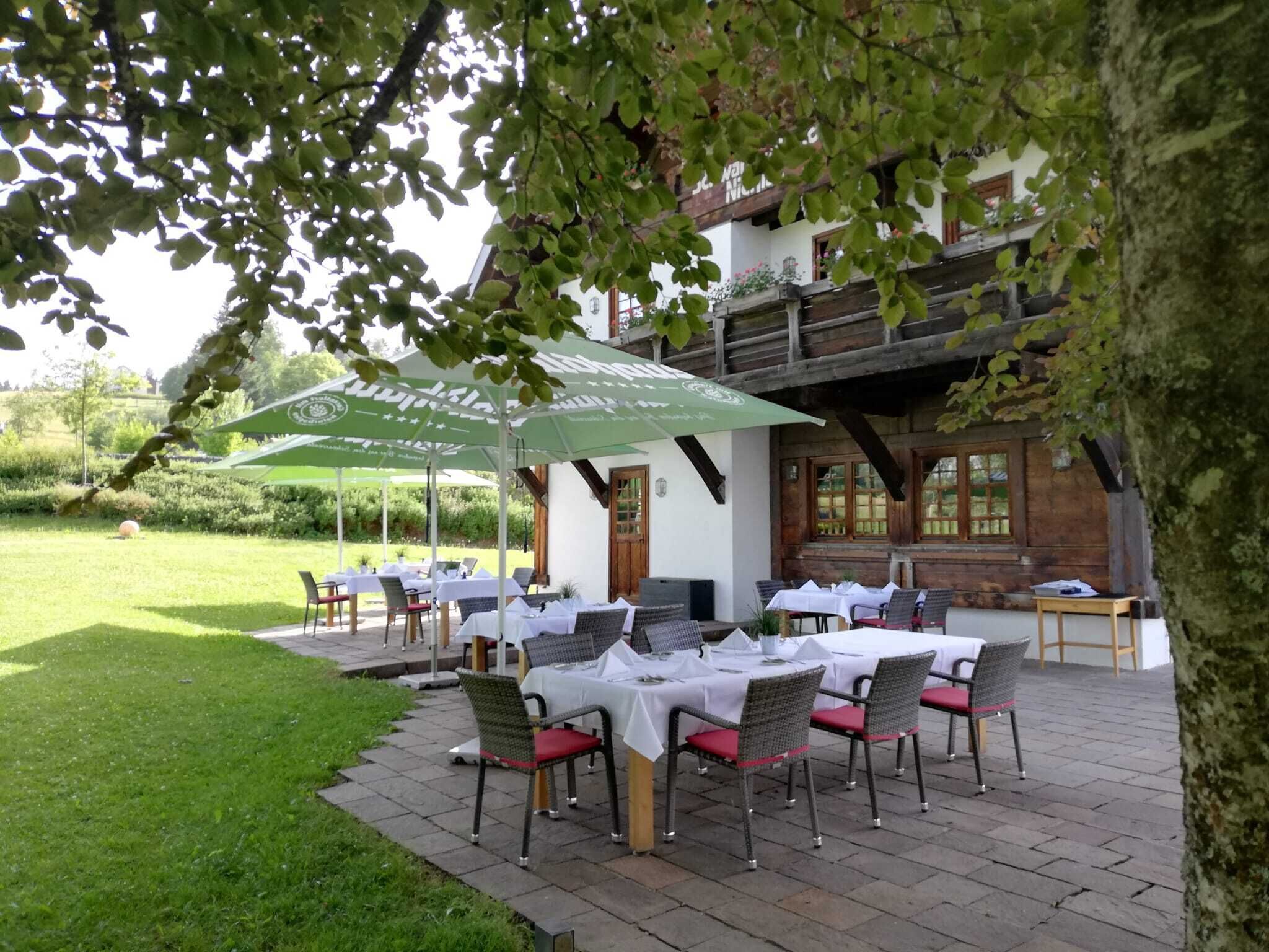 An outdoor dining area with tables and chairs, surrounded by greenery and a building with wooden balconies.