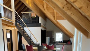 Amplia sala de estar con techo alto y vigas de madera a la vista, una escalera que sube a un nivel superior y una cocina moderna al fondo.