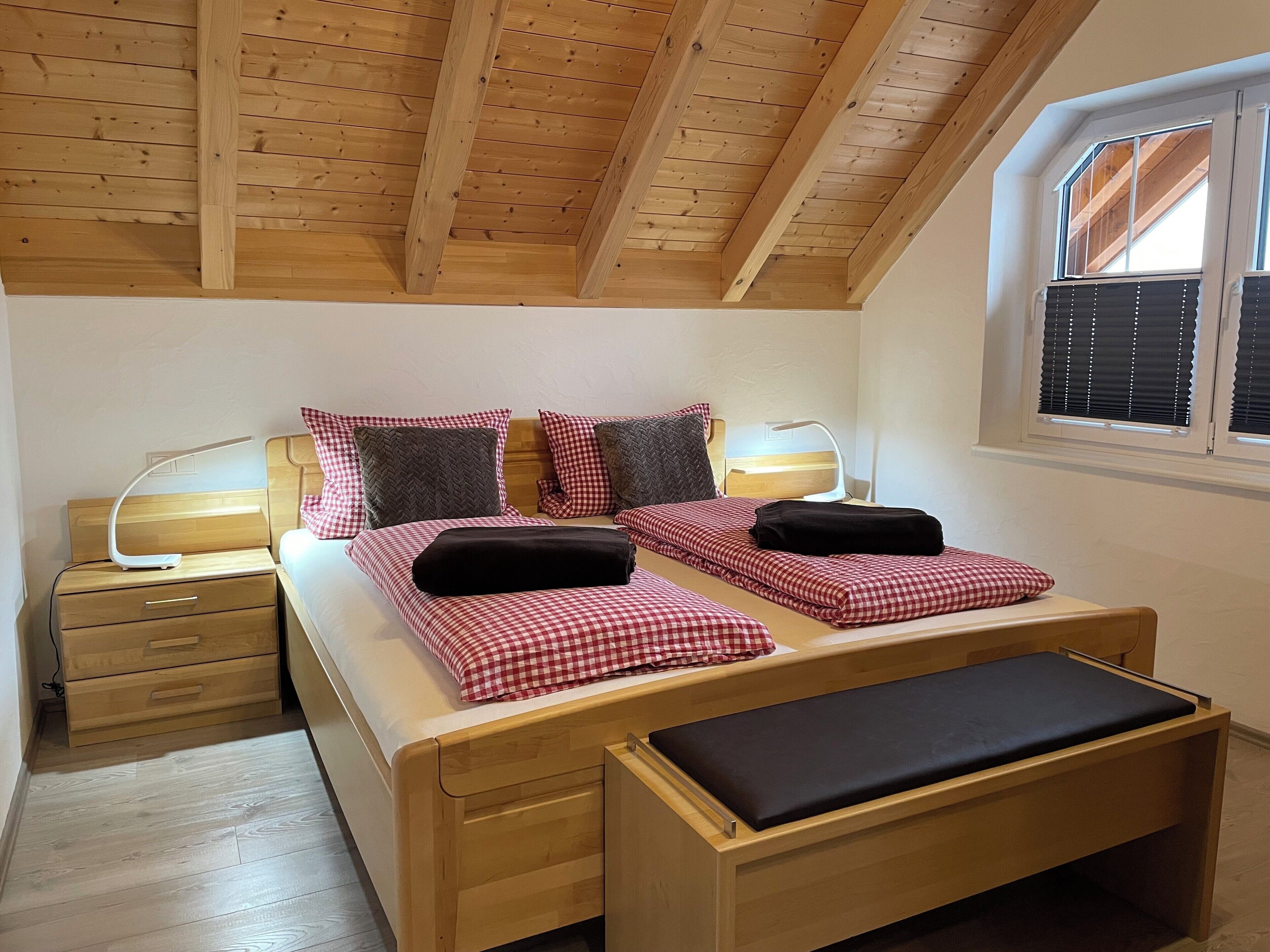 A bedroom with a wooden bed, two bedside tables, a bench, and a window with blinds.