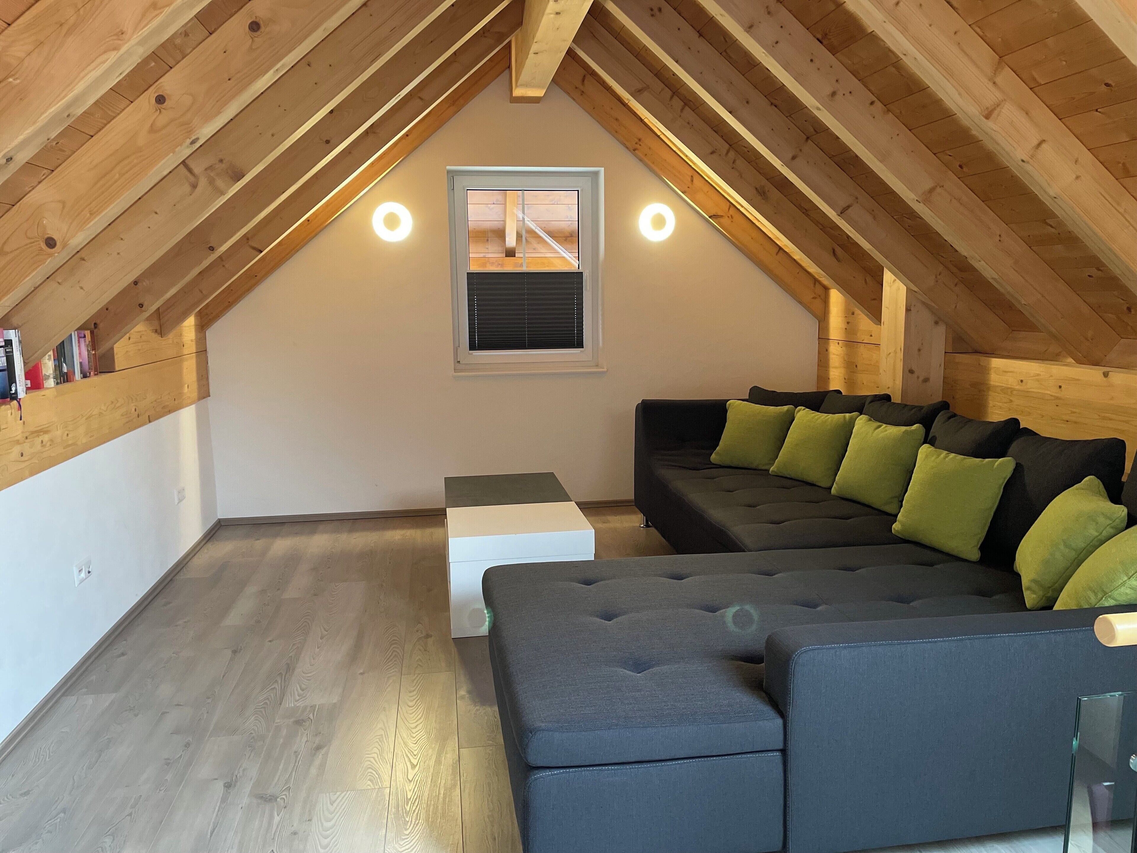 Una habitación estilo loft con techo inclinado, un sofá negro con cojines verdes, un reposapiés blanco y una ventana con persianas.