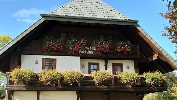 Un edificio tradicional de madera con techo inclinado, varias ventanas y un área de asientos al aire libre con mesas y sillas.