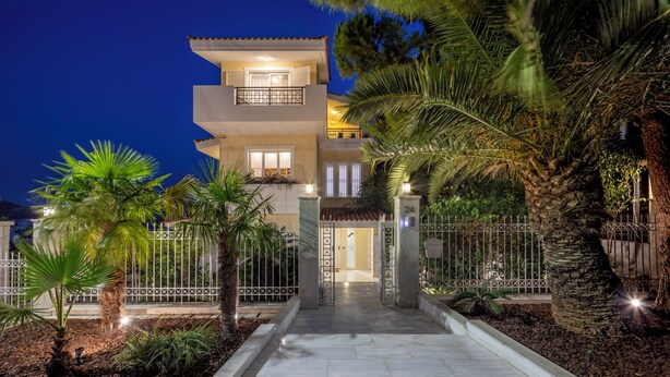 A multi-story building with a well-lit entrance, surrounded by a fence and palm trees. - The Southern Mansion (Vari-Voula-Vouliagmeni)