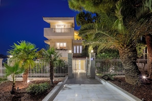 A multi-story building with a well-lit entrance, surrounded by a fence and palm trees. - The Southern Mansion (Vari-Voula-Vouliagmeni)