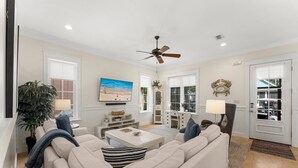 Ein geräumiges Wohnzimmer mit einem beigefarbenen Sofa, einem Couchtisch und einem Fernseher. Es gibt einen Deckenventilator, einen Kamin und ein großes Fenster.