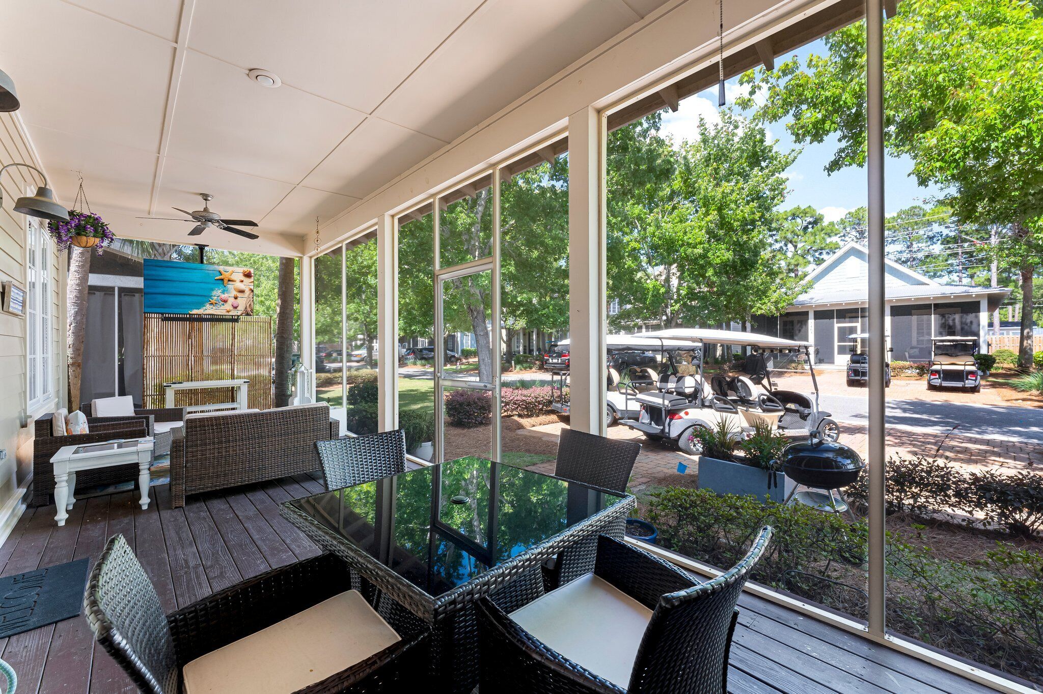 A covered outdoor patio with wicker furniture and a glass table, overlooking a street with parked cars and a building in the background.