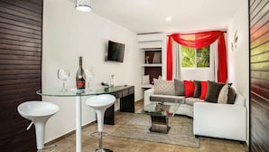 A modern living room with a glass dining table, white bar stools, a sofa with patterned cushions, and a TV mounted on the wall. - The Diamond Suites at Ahnvee (Sosúa)