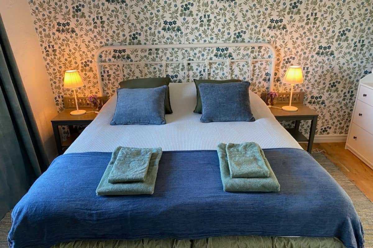 A neatly made bed with a blue and green bedspread, two pillows, and two folded towels. There are two bedside tables with lamps, and a nightstand with a lamp. The room has floral wallpaper.
