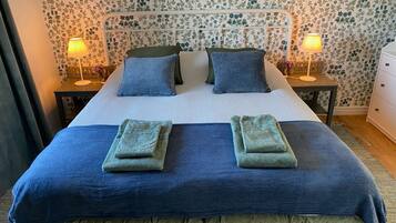 A neatly made bed with a blue and green bedspread, two pillows, and two folded towels. There are two bedside tables with lamps, and a nightstand with a lamp. The room has floral wallpaper.