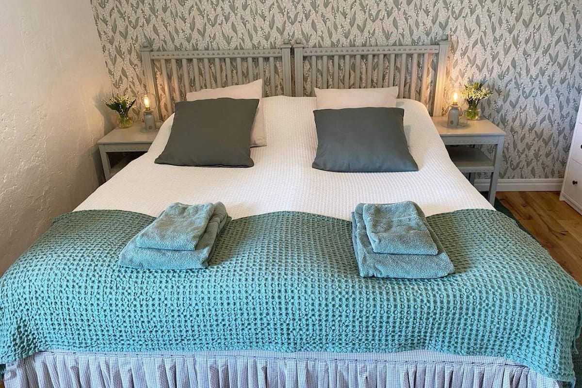 A neatly made bed with a blue knitted blanket and pillows, two bedside tables with candles, and a patterned wallpaper.