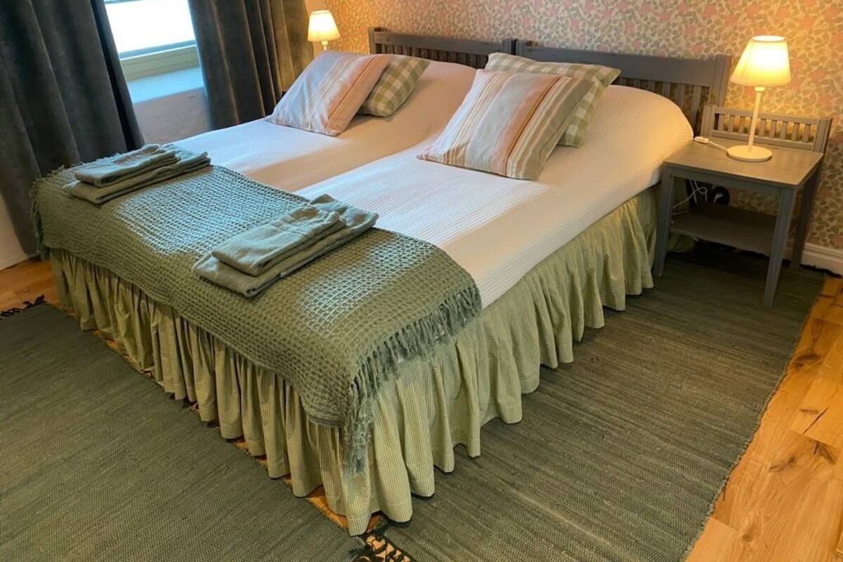 A double bed with a green bed skirt, two pillows, a bedside table with a lamp, and a patterned wall.