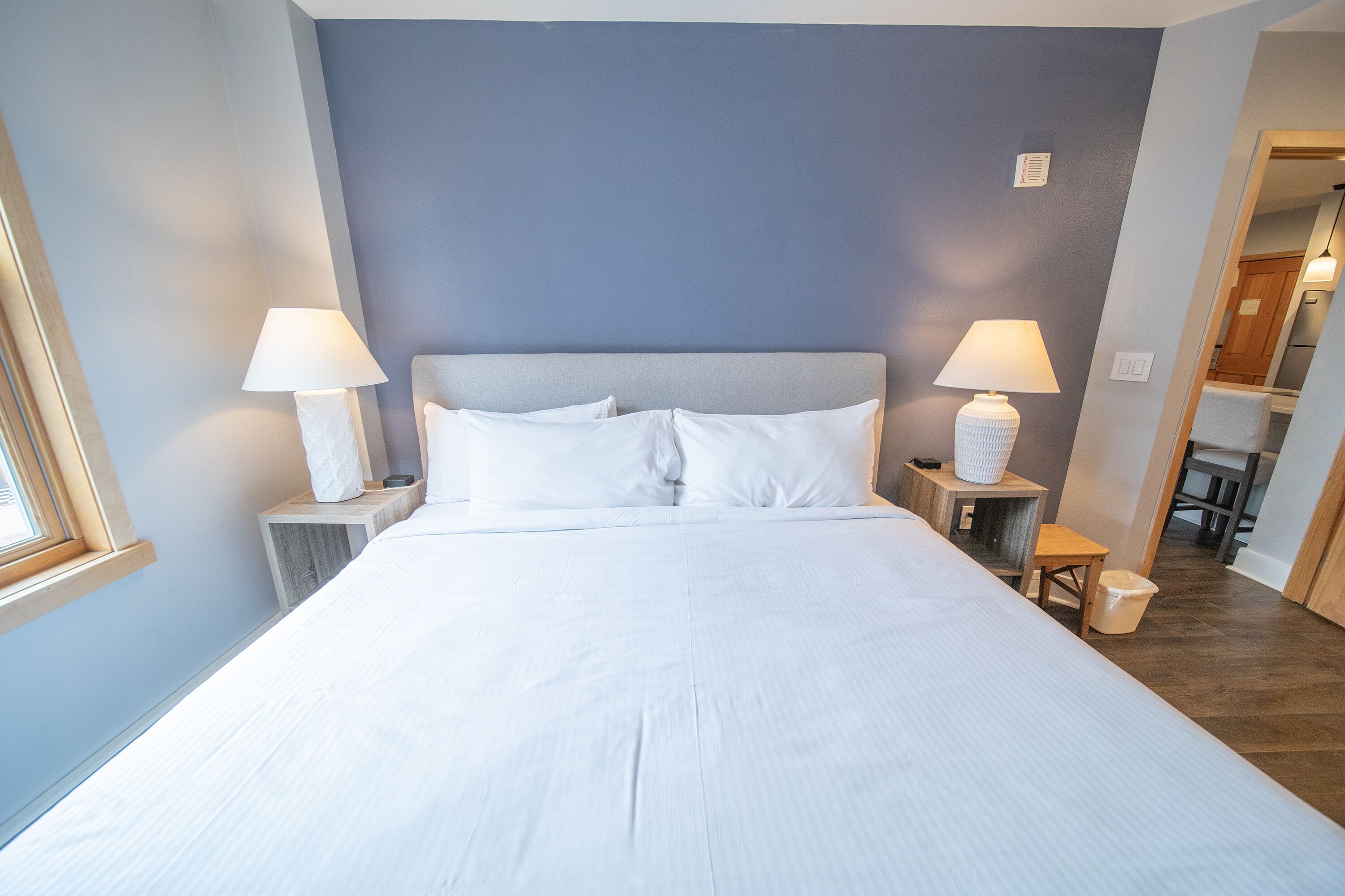 Cama bem arrumada com lençóis brancos, duas luminárias de cabeceira e uma pequena mesa de cabeceira de madeira com uma luminária.