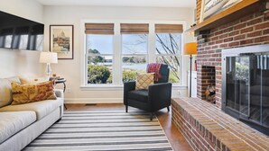 Una sala de estar con sofá, sillón, chimenea y ventanas amplias con vistas al paisaje.
