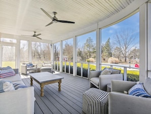 A covered outdoor deck with seating, ceiling fans, and large windows offering views of a landscaped area.