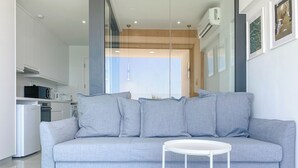 Un salon moderne avec un canapé gris, une petite table d’appoint blanche et un coin cuisine à l’arrière-plan.