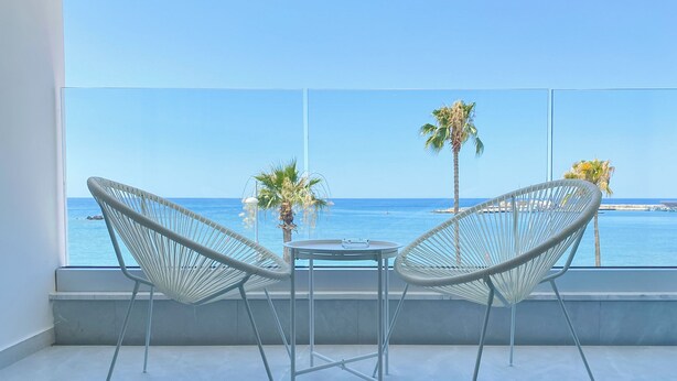 Un balcon avec deux fauteuils en osier blancs et une petite table, d’où l’on peut admirer une mer bleue et limpide, des palmiers et une côte lointaine.