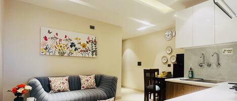 A modern living room with a sofa, a dining area with chairs, a kitchen with a sink and cabinets, and a wall clock.