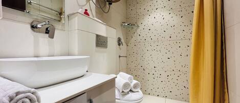 Un baño con lavamanos blanco, inodoro y ducha. Hay toallas en el inodoro y un cabezal de ducha sobre la ducha.