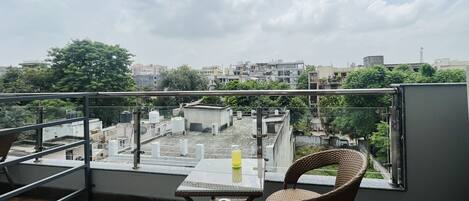 Un balcón con una mesa y sillas de mimbre, con vistas a un paisaje urbano con edificios y árboles.