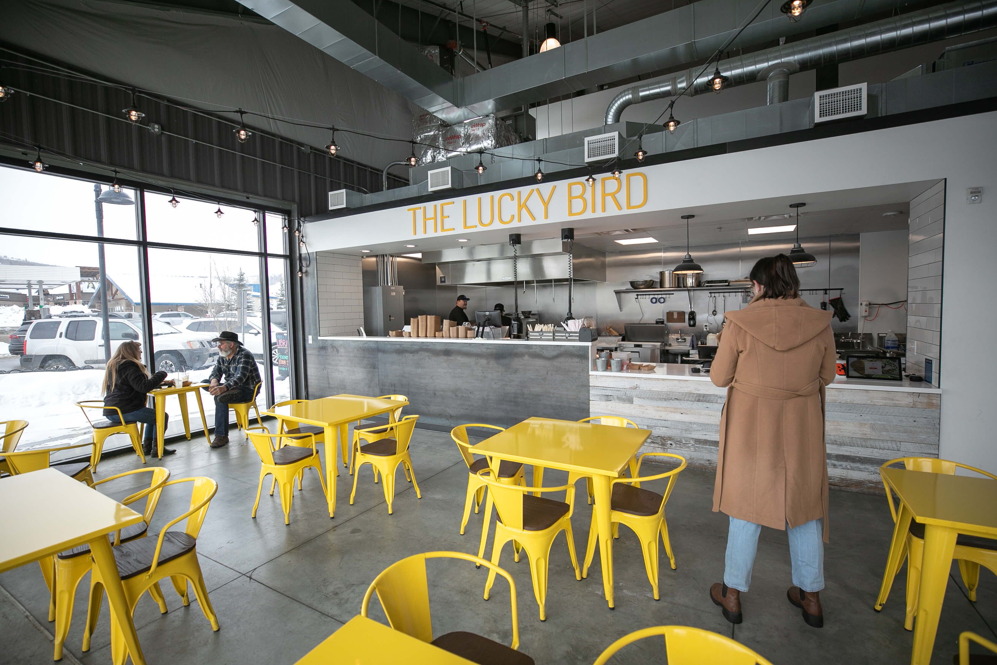 Un área de estar interior con mesas y sillas amarillas, una zona de cocina con personal, y un letrero que dice THE LUCKY BIRD.