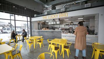 Un área de estar interior con mesas y sillas amarillas, una zona de cocina con personal, y un letrero que dice THE LUCKY BIRD.
