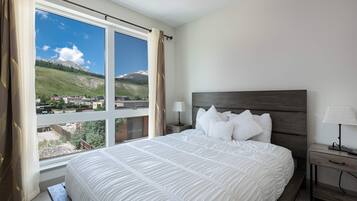 Un dormitorio con una cama grande, una mesita de noche, una lámpara y vistas a un paisaje montañoso a través de una ventana.