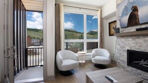 A modern living room with a fireplace, two white armchairs, a small table, and a large window offering a mountain view. - Fourth Street Crossing Traverse Townhome: Stylish Retreat (Silverthorne)