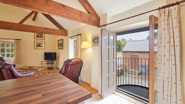 A cozy room with a wooden ceiling, a dining table, leather chairs, a TV, and a large window with a view of a brick building outside. - Host & Stay | Widger Mews Apartment (Malton)