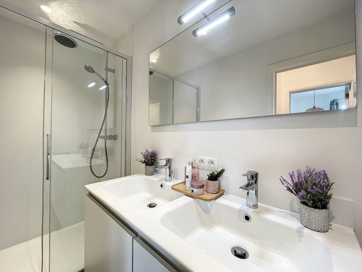 Een moderne badkamer met een douchecabine van glas, een wit wastafelmeubel met een houten dienblad en twee potplanten.