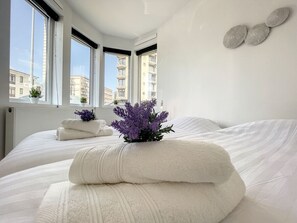 Une chambre d’hôtel avec un grand lit, une fenêtre donnant sur des immeubles et une petite plante posée sur le lit.