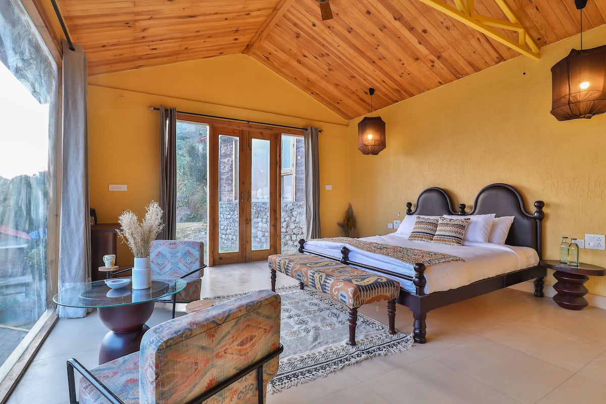 Une chambre avec un grand lit, une table en verre, un banc et une vue à travers la porte vitrée.