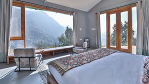 A bedroom with a large bed, a chair, a bench, and a view of mountains through large windows. - Seclude Pangot Perch (Nainital)