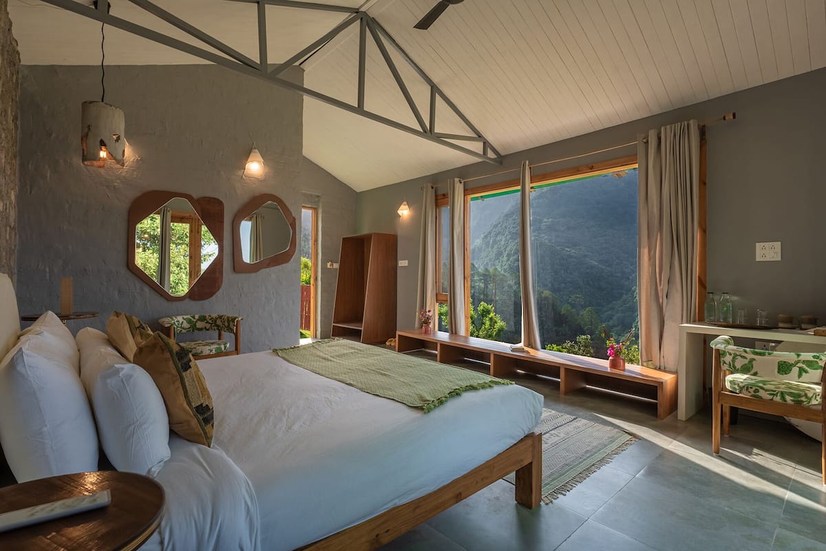 Une chambre avec un grand lit, une tête de lit en bois, deux miroirs fixés au mur, un banc en bois, une table de chevet et une vue sur une forêt, visible à travers de grandes fenêtres.