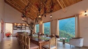 A dining area with a wooden table, chairs, and a large window with a view of a forested mountain. - Seclude Pangot Perch (Nainital)